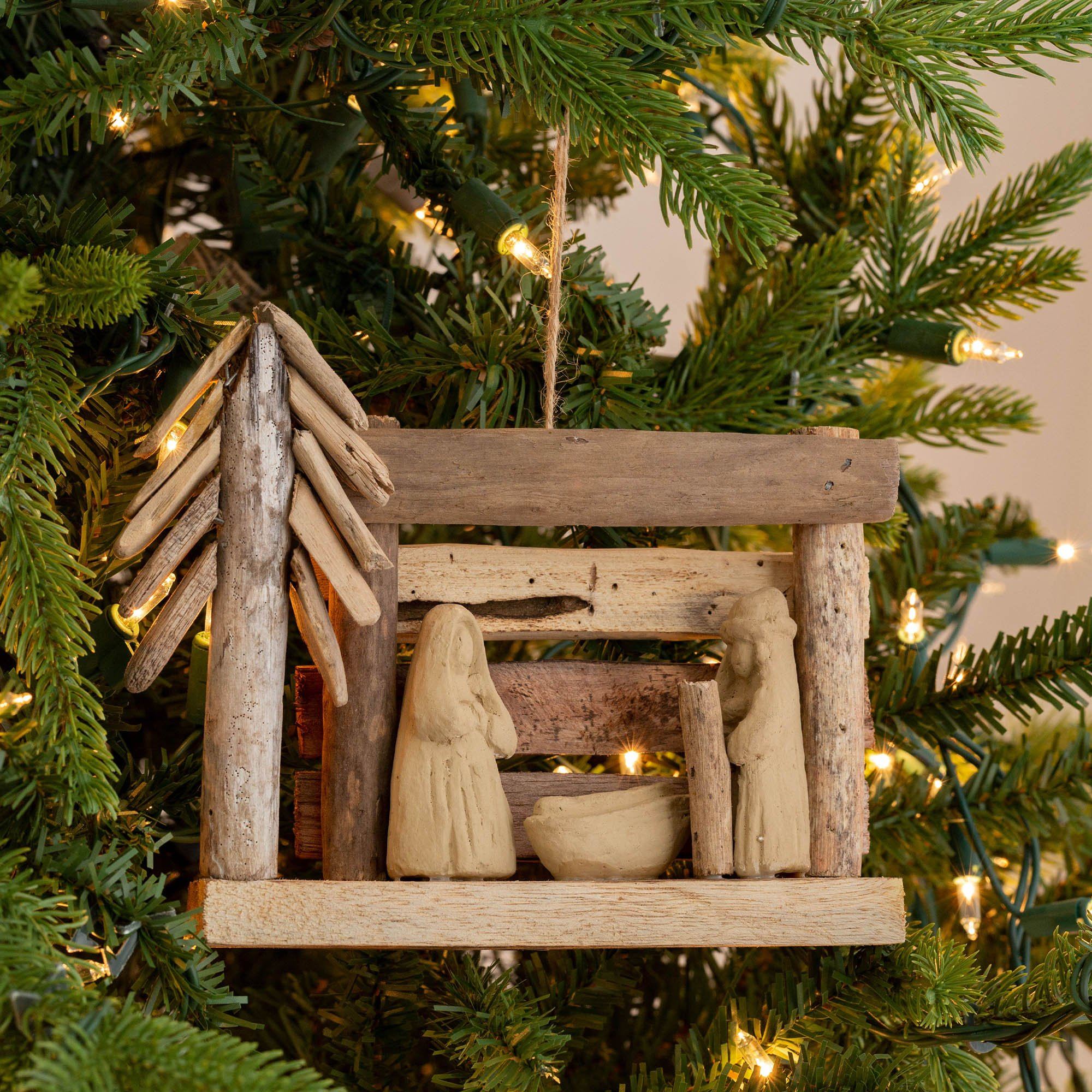 Recycled driftwood nativity ornament with handmade clay figures, hanging on a Christmas tree with warm lights.