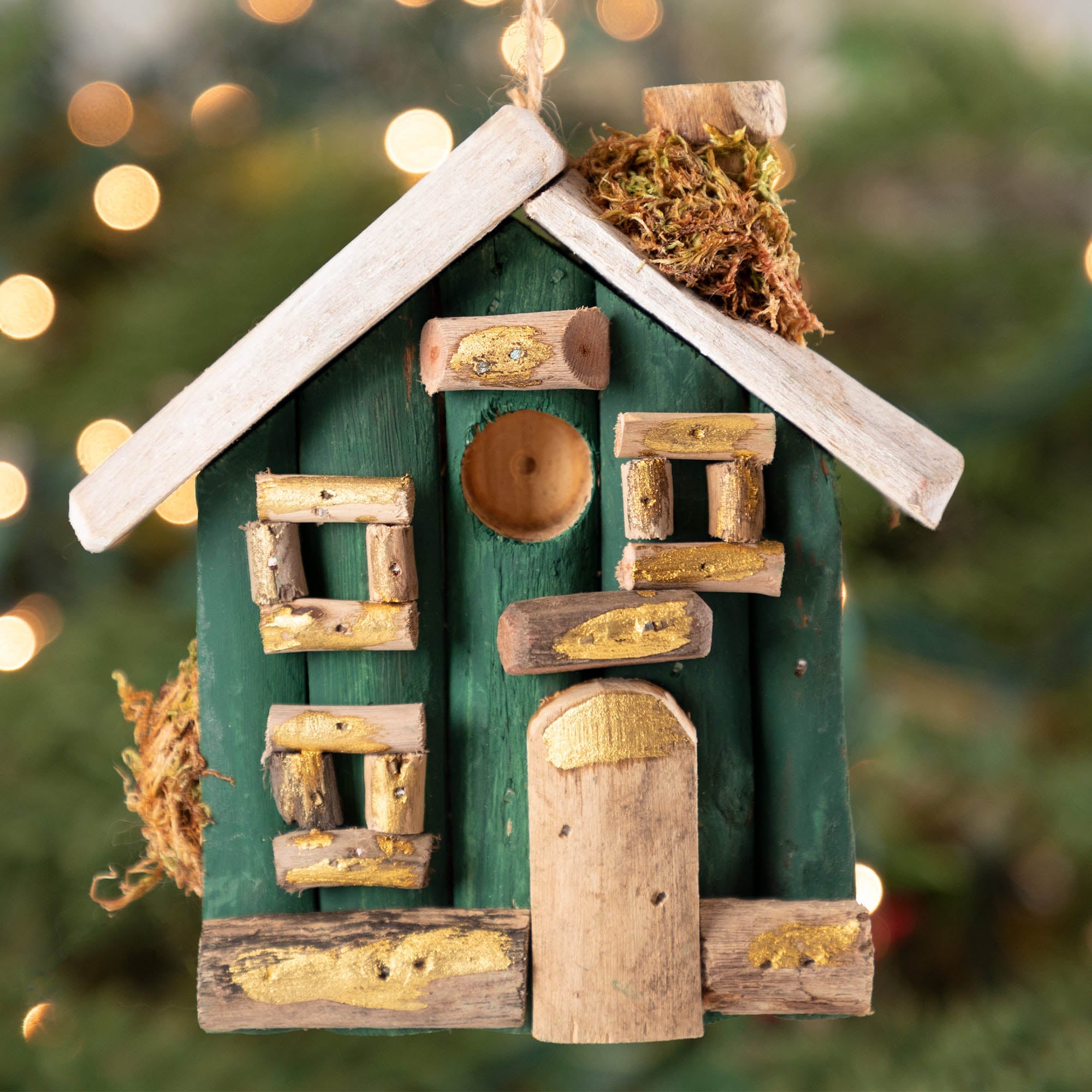 Close view of a hand holding a handcrafted driftwood Christmas house ornament with festive green paint. Eco-friendly and fair-trade, made from recycled driftwood.