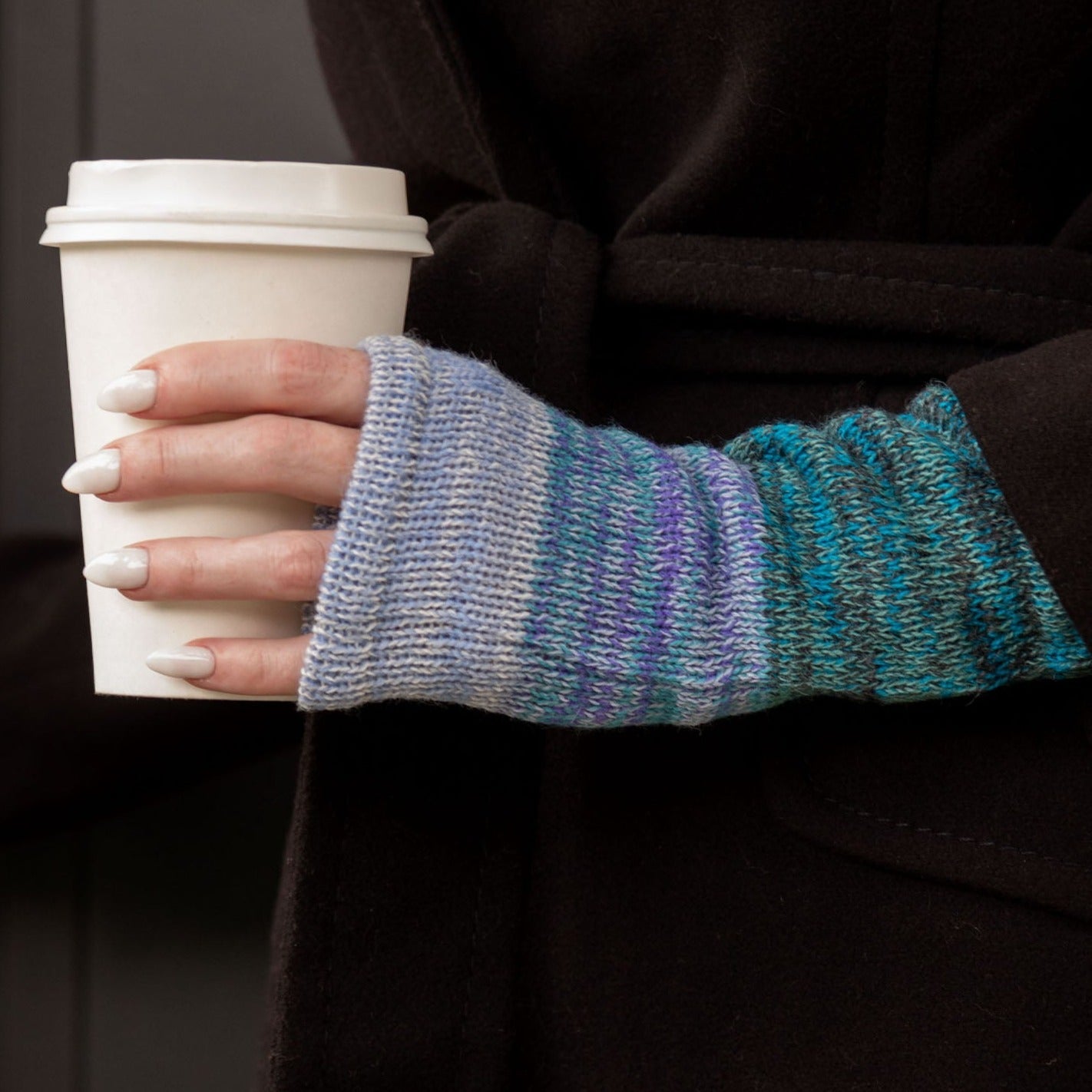 A hand wearing a colorful handmade alpaca fingerless mitten holding a paper cup filled with coffee indoors.