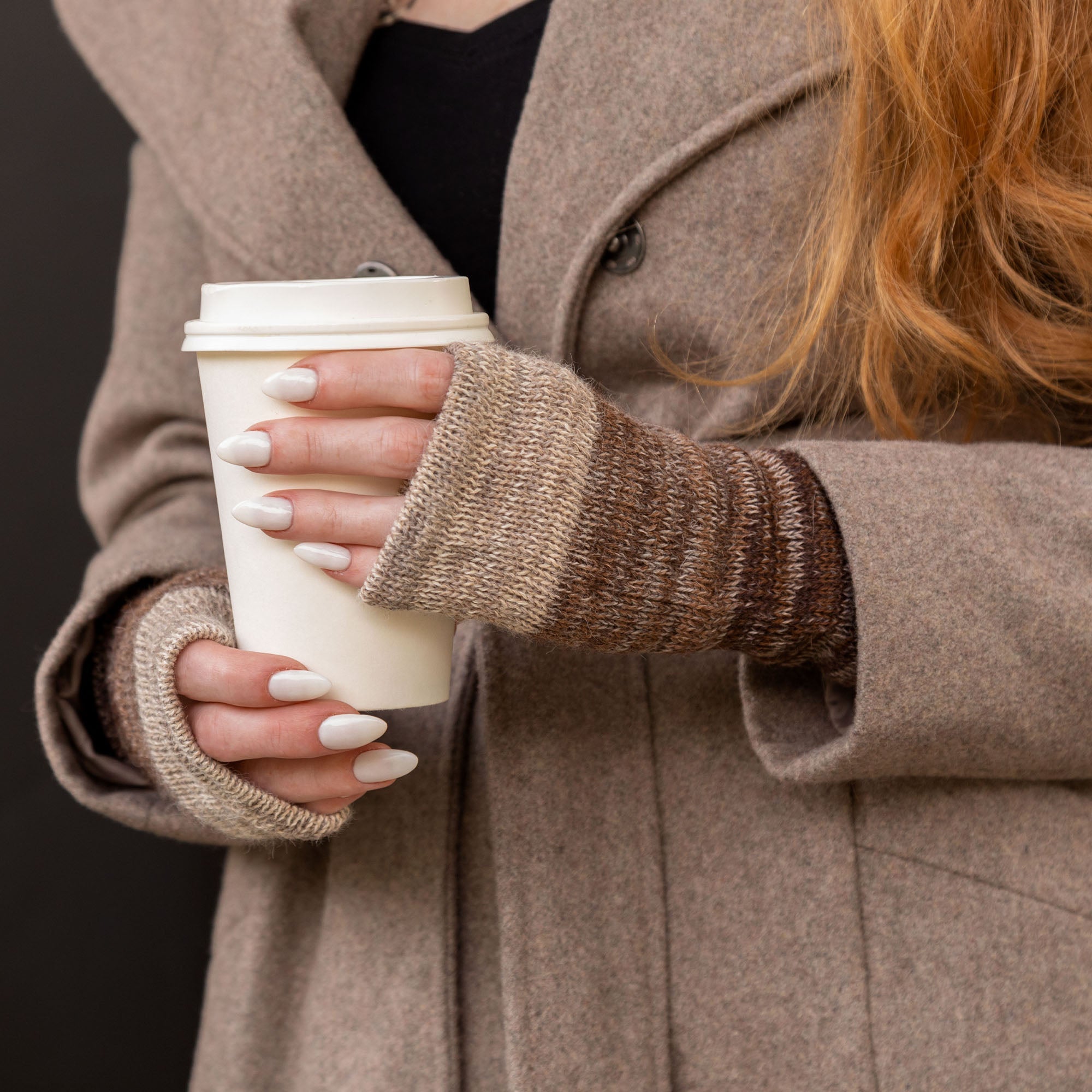 Hands holding a paper cup while wearing striped alpaca fingerless mittens in warm brown tones. Handmade in Bolivia from 100% alpaca wool.