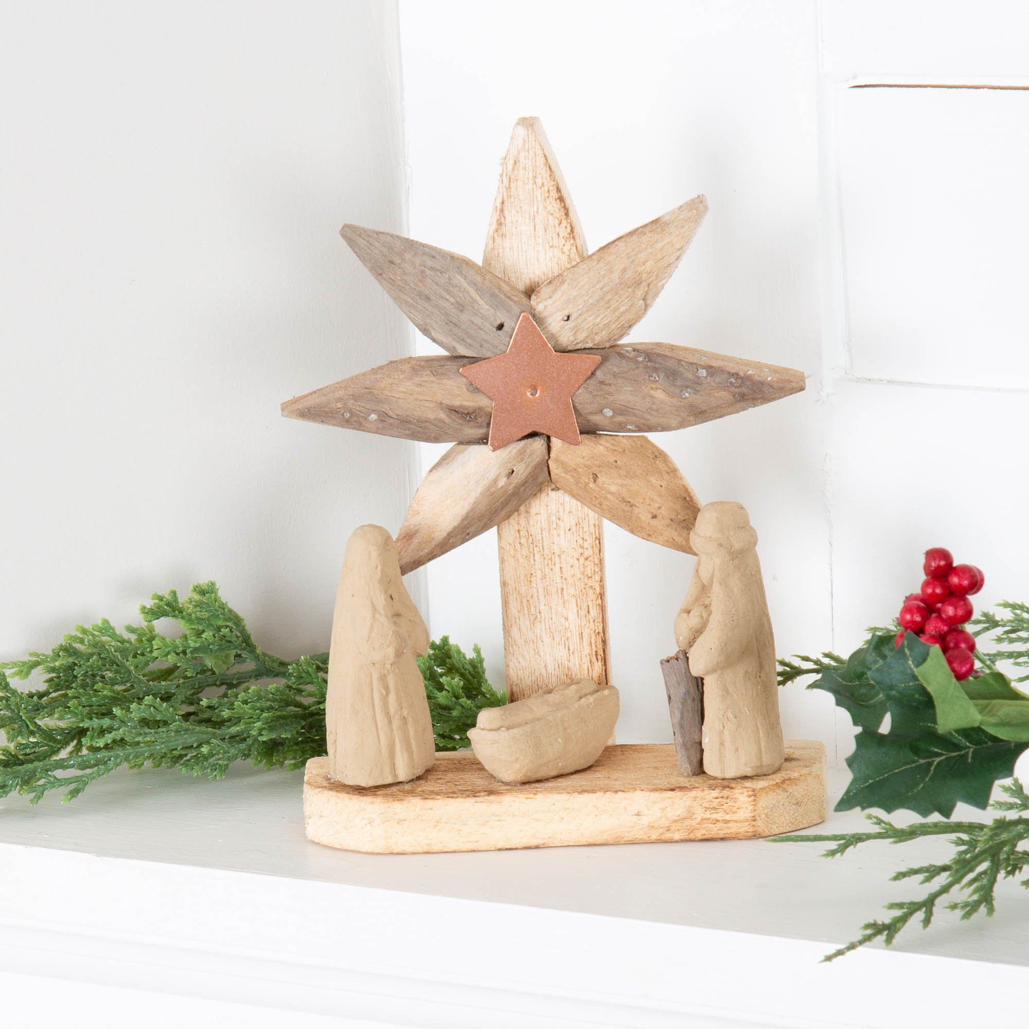 Recycled, handmade driftwood star decoration with clay nativity figures, copper star center, and festive greenery on a white mantel.