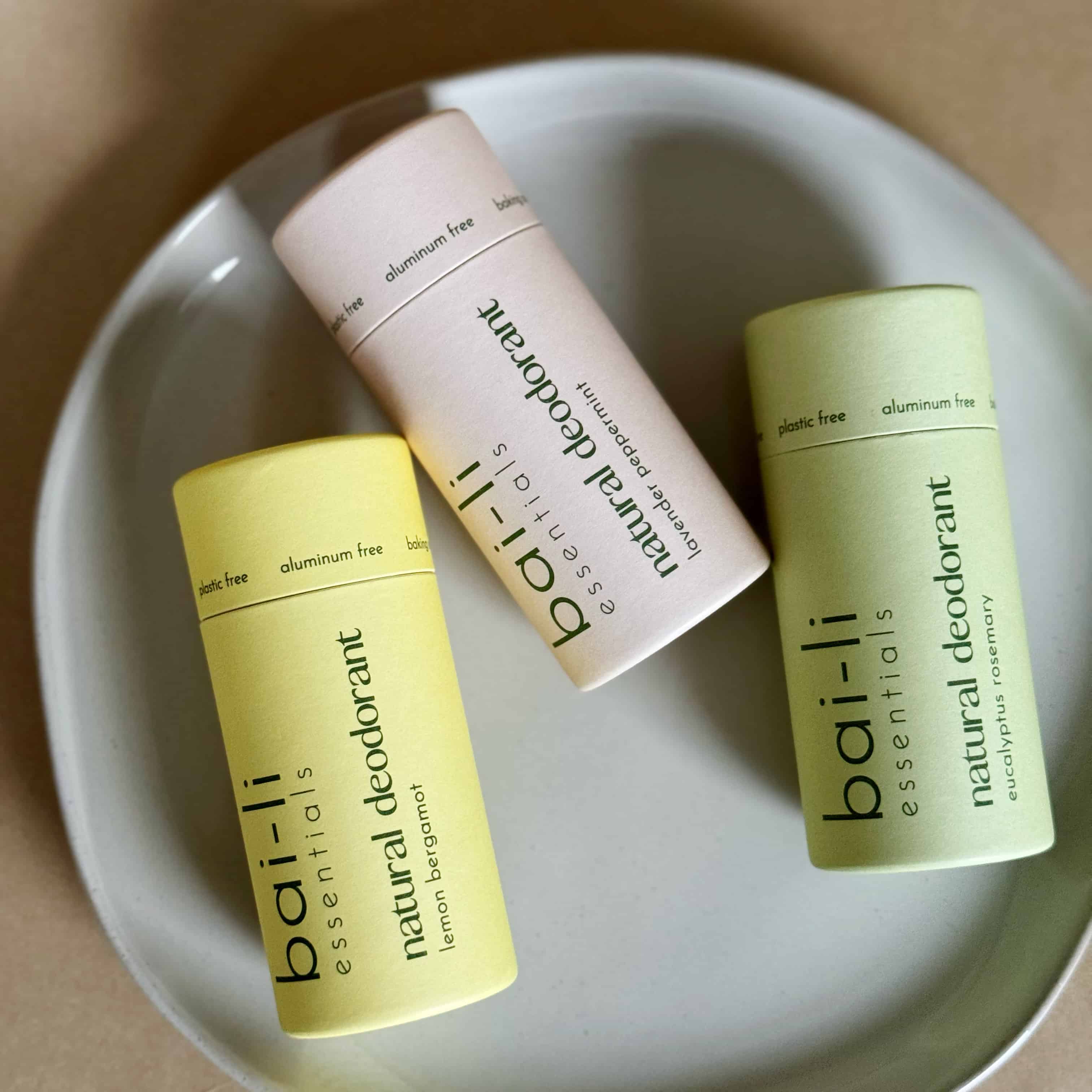 Three plastic-free deodorant tubes—Lavender + Peppermint, Cedarwood + Sweet Orange, and Lemon + Bergamot—arranged on a gray ceramic dish.