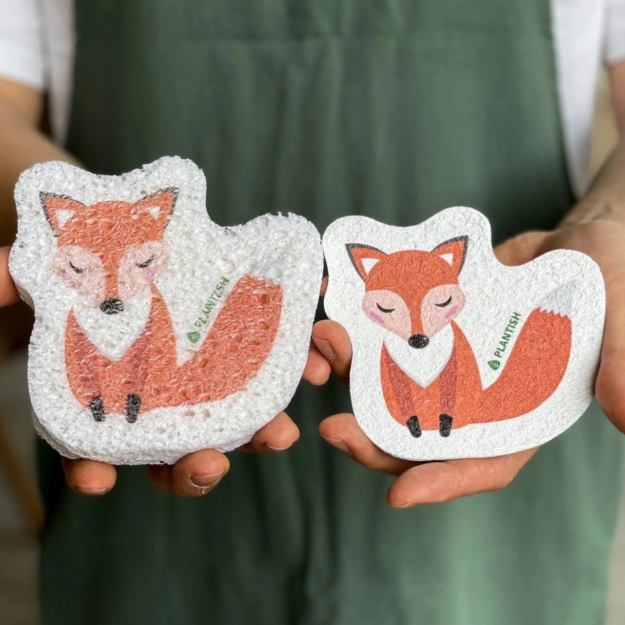 A person holds two fox-shaped sponges against a green background. One sponge is a compressed, flat shape, while the other is an expanded, porous shape. Both sponges are labeled PLANTISH.