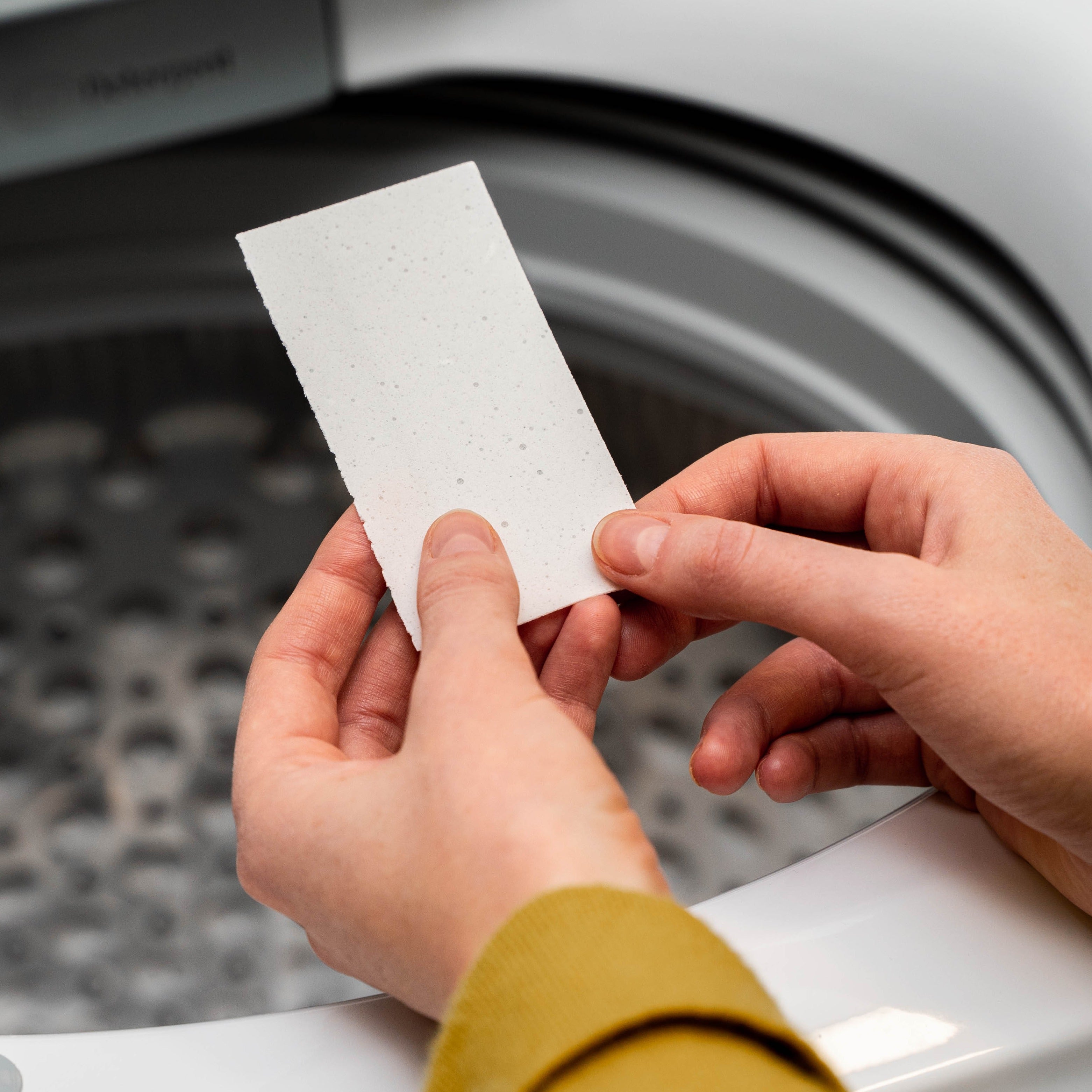 Lifestyle shot of a washing machine door partially open with a person’s hand loading white laundry detergent strips inside.