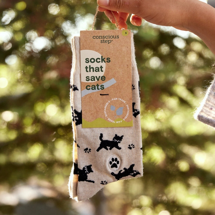 A hand holds a pair of Conscious Step socks with a beige background and a pattern of black cats and paw prints. The tag identifies them as "socks that save cats" and notes they are sustainable, fair trade and organic.