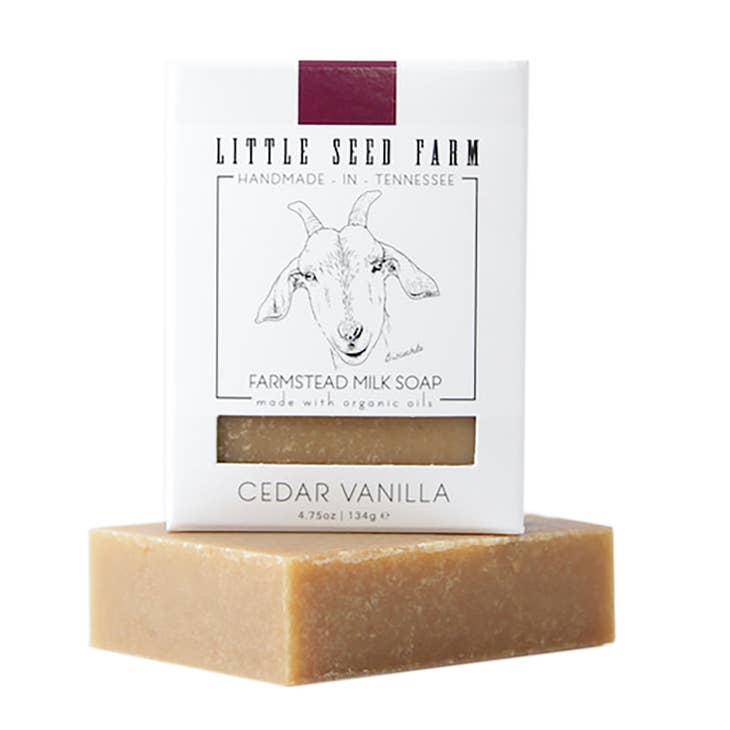 A bar of Little Seed Farm soap sits underneath its white cardboard packaging. The soap is a light golden brown color. The front of the box features a line drawing of a goat and the text: Farmstead Milk Soap Cedar Vanilla is printed below it.
