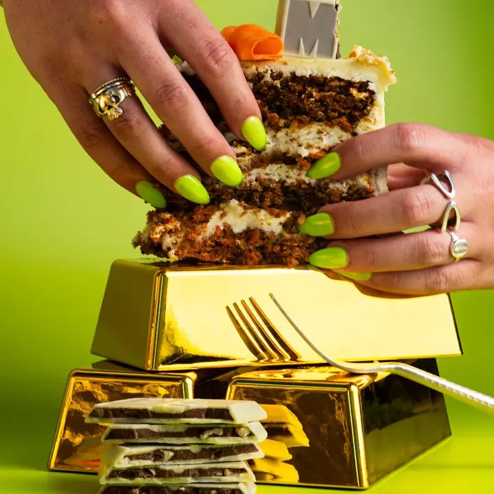 Two hands holding a slice of carrot cake topped with frosting, visually evoking the flavor of the Maeve “14-Carrot Cake” truffle bar.