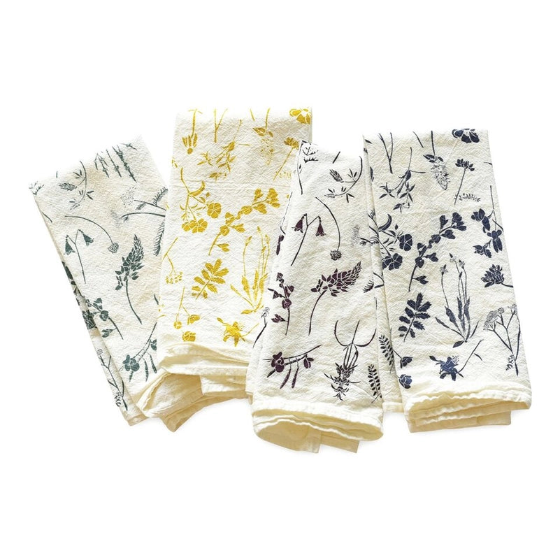 A sweet set of four white napkins is shown folded, each with a charming botanical pattern in a single color: dusty green, yellow, dark brown and deep blue.