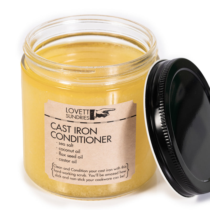 An open glass jar of "Lovett Sundries Cast Iron Conditioner" with a black lid next to it. The label lists the ingredients as: sea salt, coconut oil, flax seed oil and castor oil.