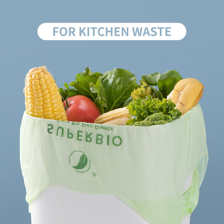 A pale green compostable bag filled with kitchen waste like a corn cob a tomato and leafy vegetables. A white oval banner at the top of the image reads: "FOR KITCHEN WASTE".