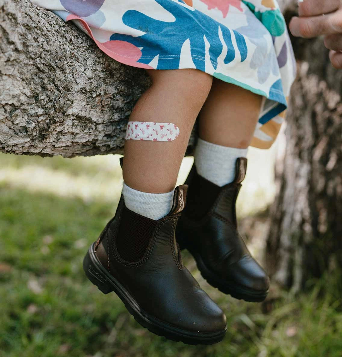 A child's legs dangle from a tree branch outdoors, wearing colorful socks and boots, with a unicorn-print bamboo bandage on one knee—made from breathable, non-toxic materials designed for sensitive skin.