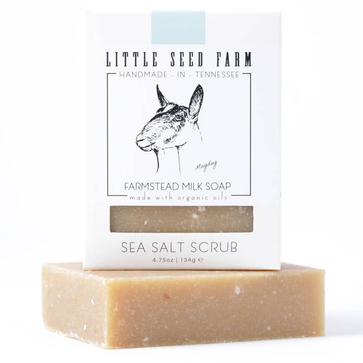 A creamy bar of Little Seed Farm soap sits on a white surface with its packaging behind it. The box shows a drawing of a goat and the words Sea Salt Scrub.