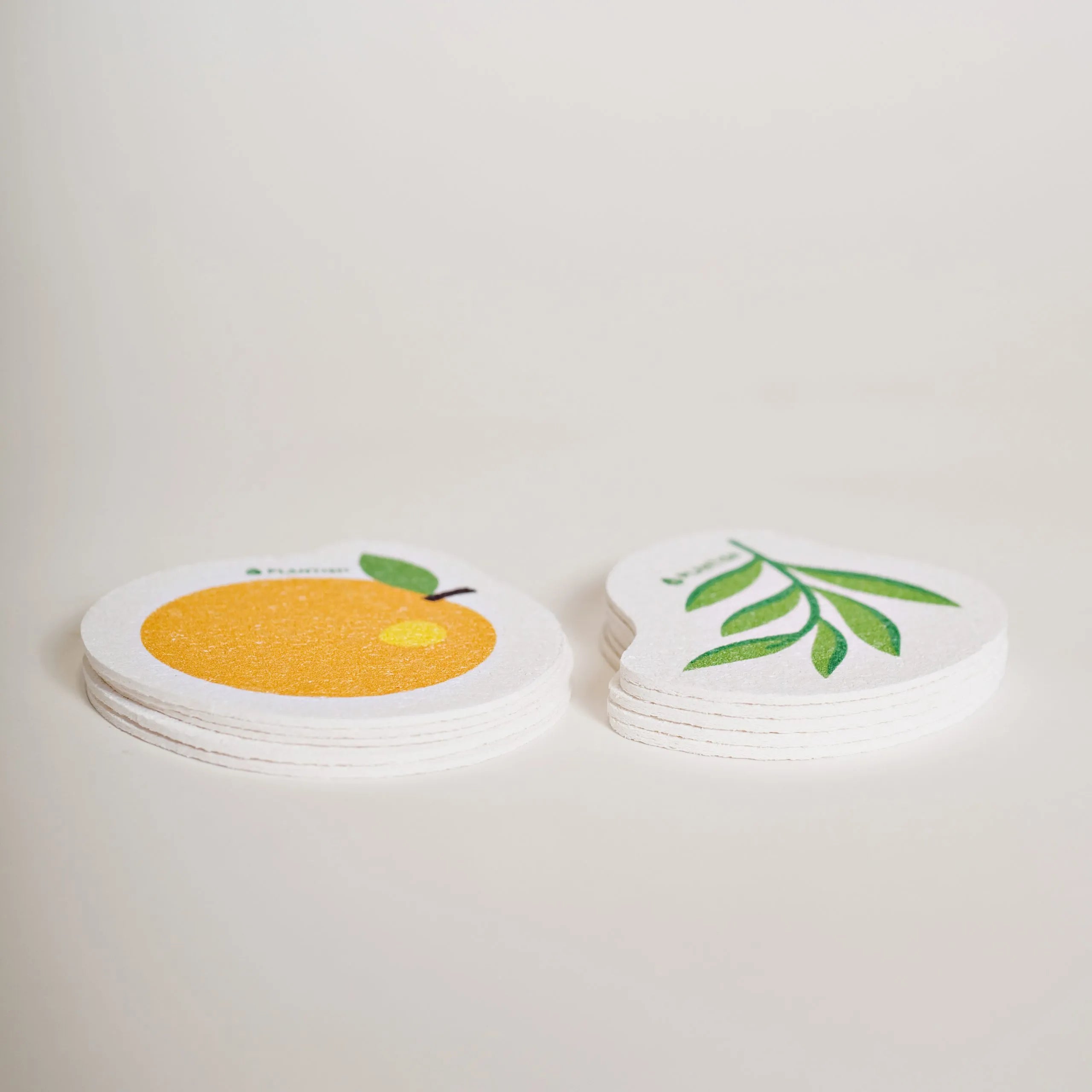 Two stacks of thin, compressed PLANTISH pop-up sponges are shown on a white background. The stack on the left is shaped and designed like an orange, while the one on the right is shaped like a leaf.