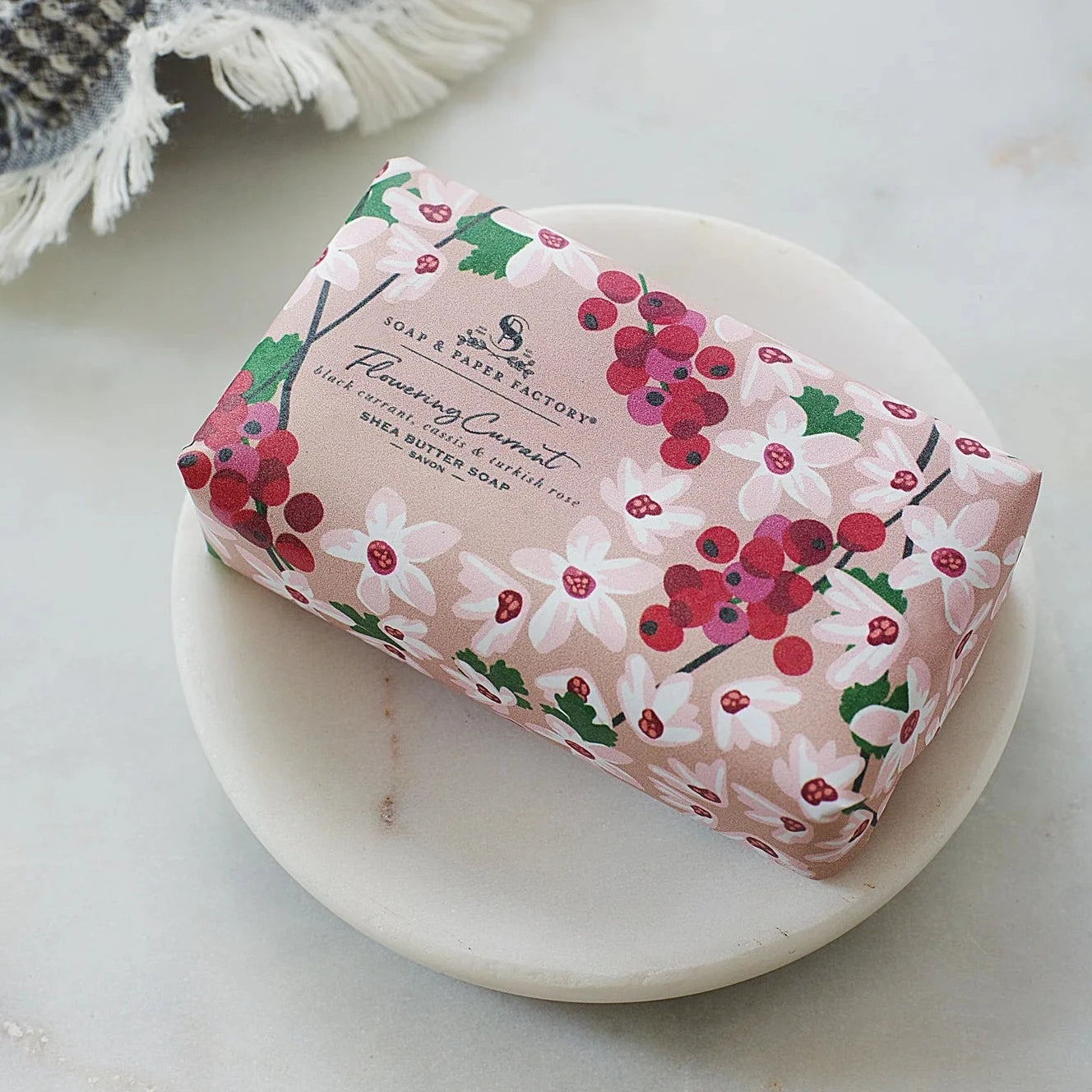 A "Flowering Currant" 'Soap & Paper Factory' shea butter soap bar rests in a small, round white dish. The soap is wrapped in pink paper with a white and red floral design.
