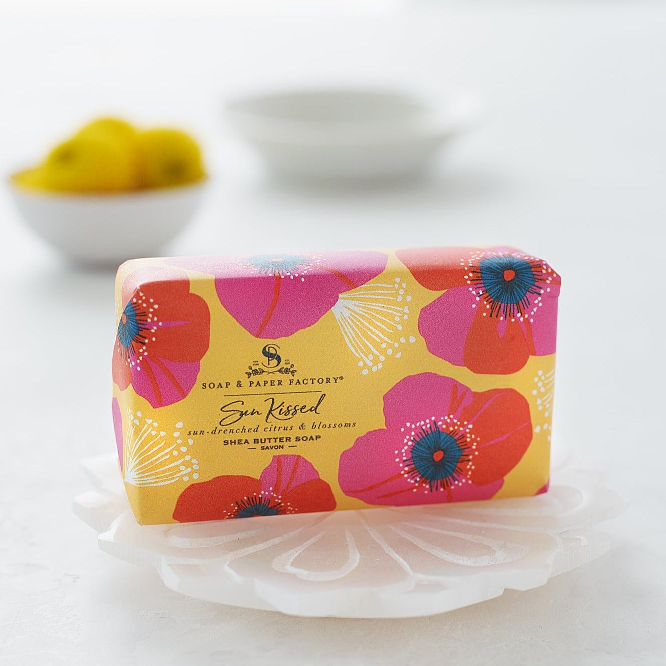 A brightly colored "Sun Kissed" shea butter soap bar sits on a small white shell-shaped dish. The soap wrapper is yellow with a pattern of large pink and orange blossoms.