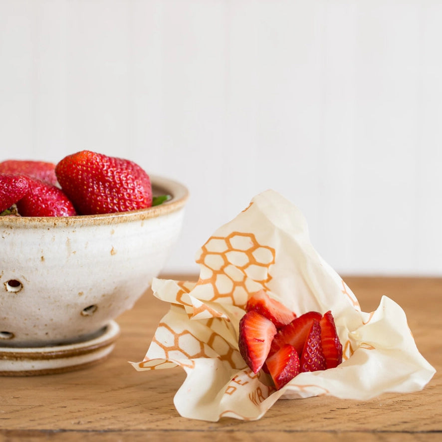 Close-up of a ceramic bowl filled with fresh strawberries beside a beeswax wrap holding sliced strawberries on a wooden surface.