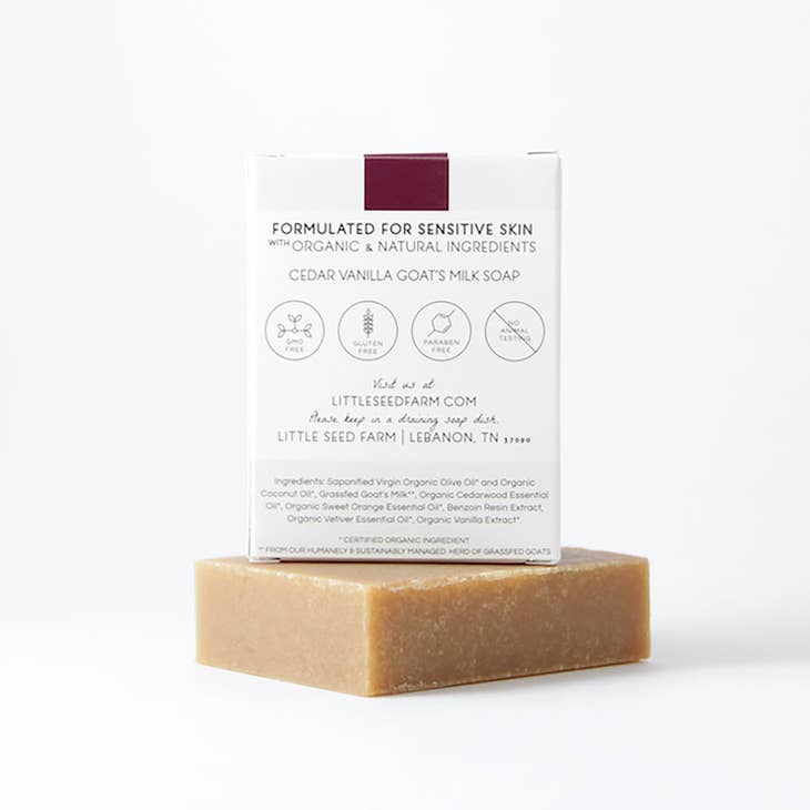 A Cedar Vanilla Goat's Milk Soap bar sits in front of its packaging which lists the ingredients and features. The soap is formulated for sensitive skin with organic ingredients and is certified cruelty-free gluten-free and paraben-free. It is made from a sustainably managed herd of goats.