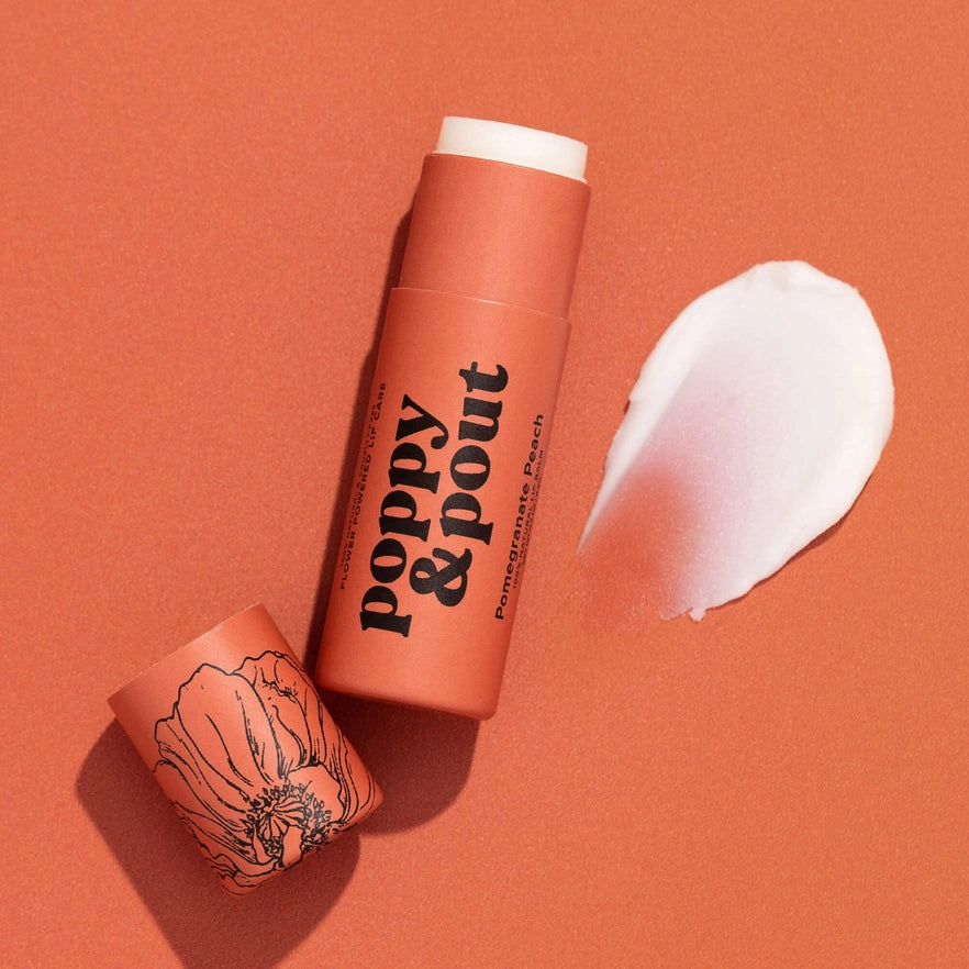 An open tube of poppy & pout lip care is shown on a reddish-orange background. The tube is labeled "Pomegranate Peach" and has a poppy flower illustration on the cap. A white product swatch with a translucent pink tint is smeared next to the tube.