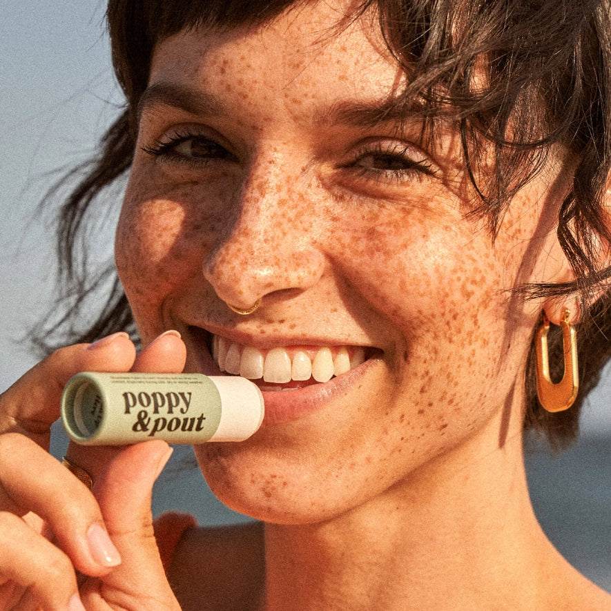 A smiling person with freckles holds a tube of poppy & pout lip care to their mouth. The lip care tube is a pale green color with the brand's name printed in white. The person is wearing a large, gold, oval earring.