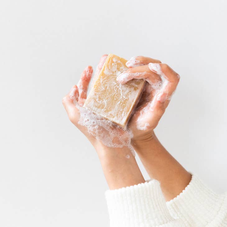 A person's hands hold a bar of golden-brown soap covered in white foam. The person is wearing a white sweater against a plain white background.
