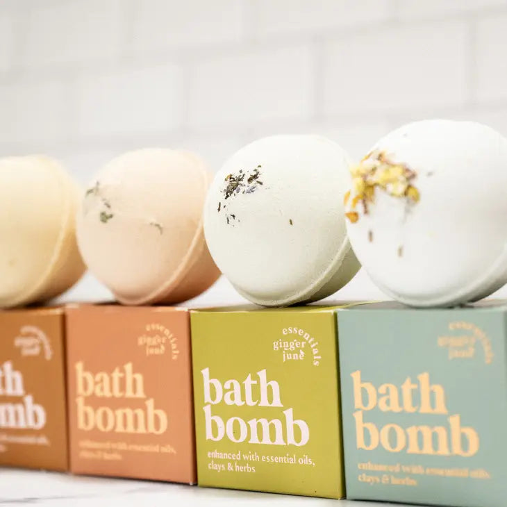 A line of four botanical bath bombs is shown, each sitting on a square box. The bath bombs and their corresponding boxes are different colors, each bath bomb is topped with various dried herbs and flowers. The boxes are labeled "ginger june essentials bath bomb".