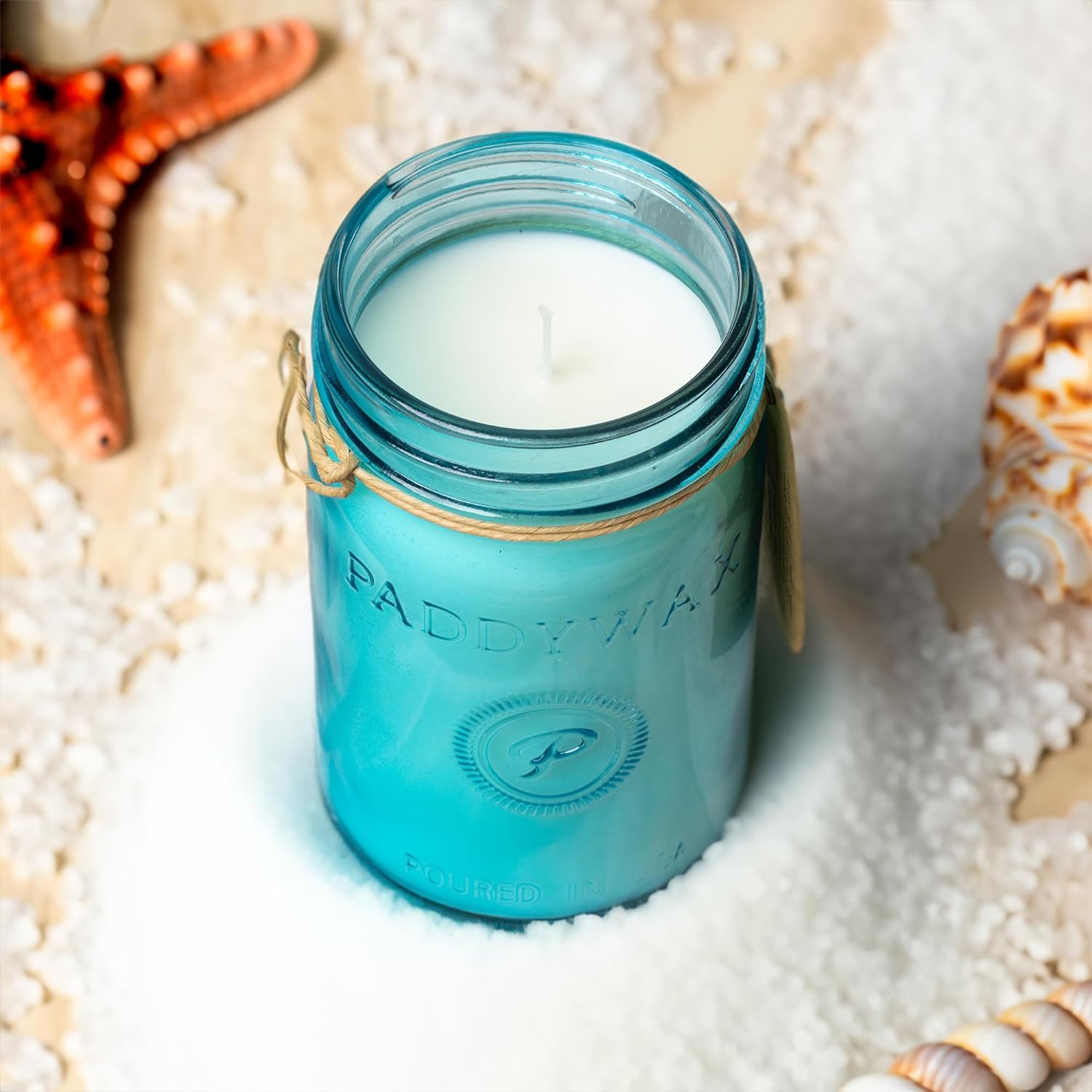 A lit blue Paddywax "Ocean Tide + Sea Salt" candle is shown in a jar. It is resting in a pile of white salt, with a starfish in the background.