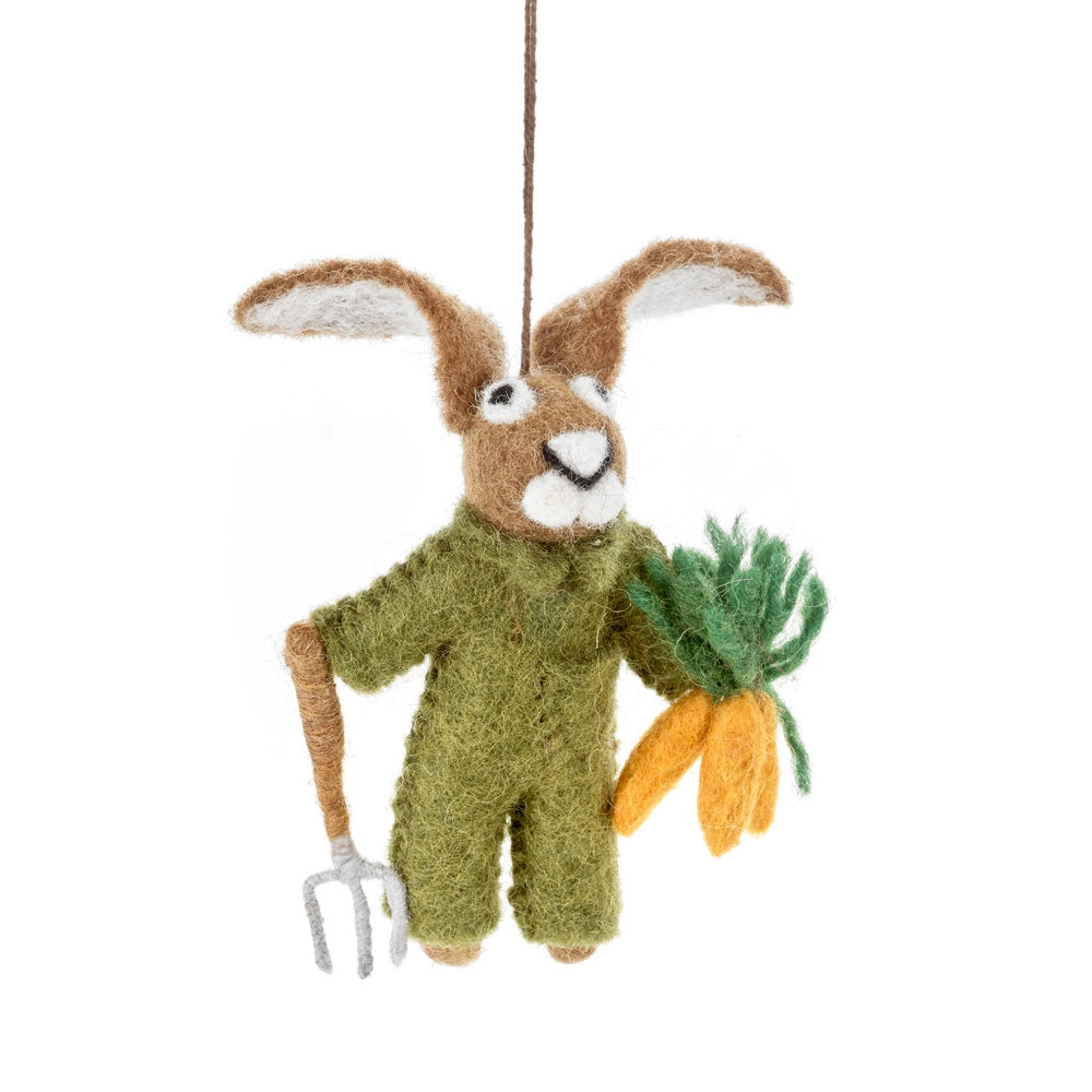 A handmade felt rabbit hanging decoration is shown, dressed in a green jumpsuit. The rabbit is holding a miniature pitchfork in one hand and a bouquet of carrots in the other.