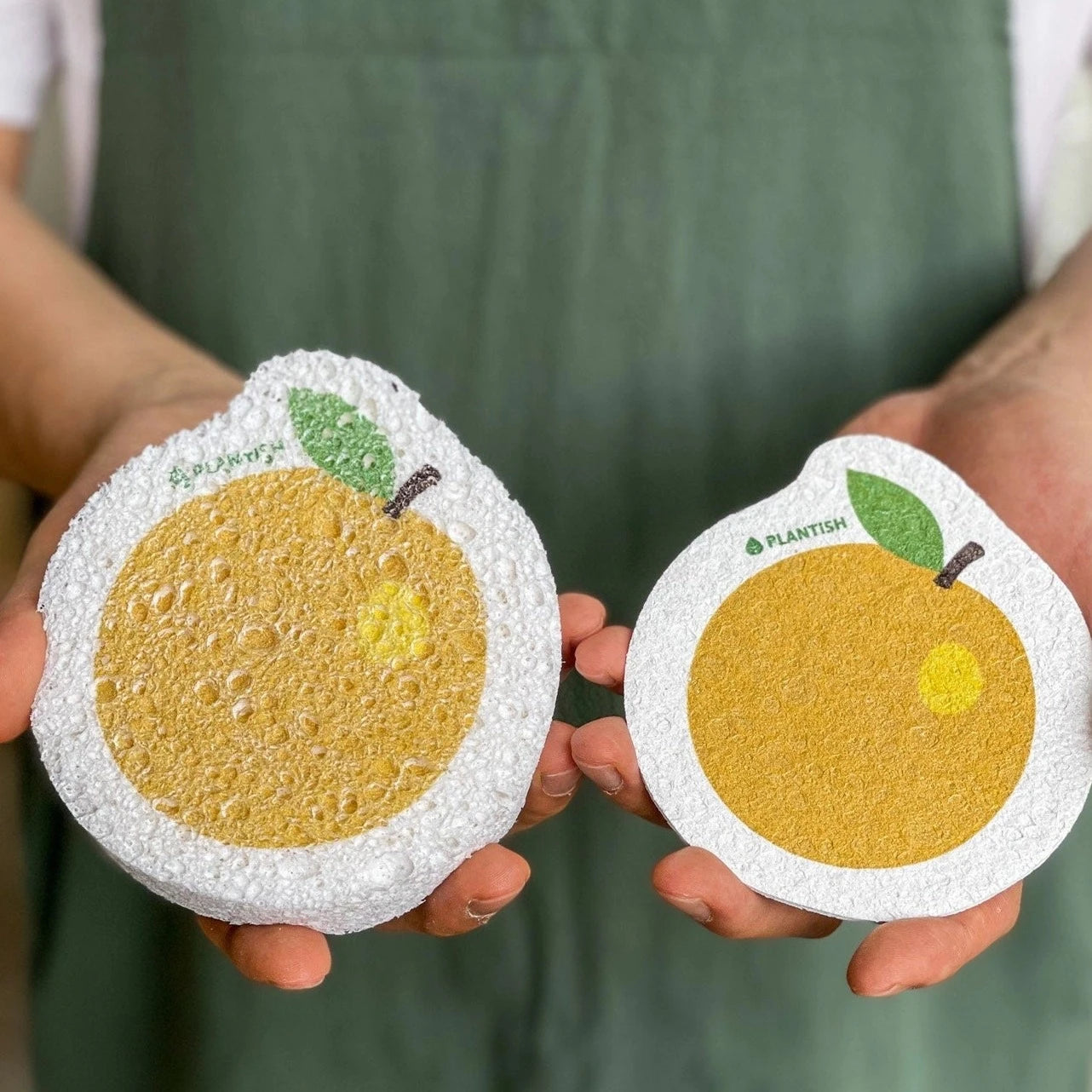 A person is holding two PLANTISH orange-slice sponges. One sponge is flat, and the other has expanded.