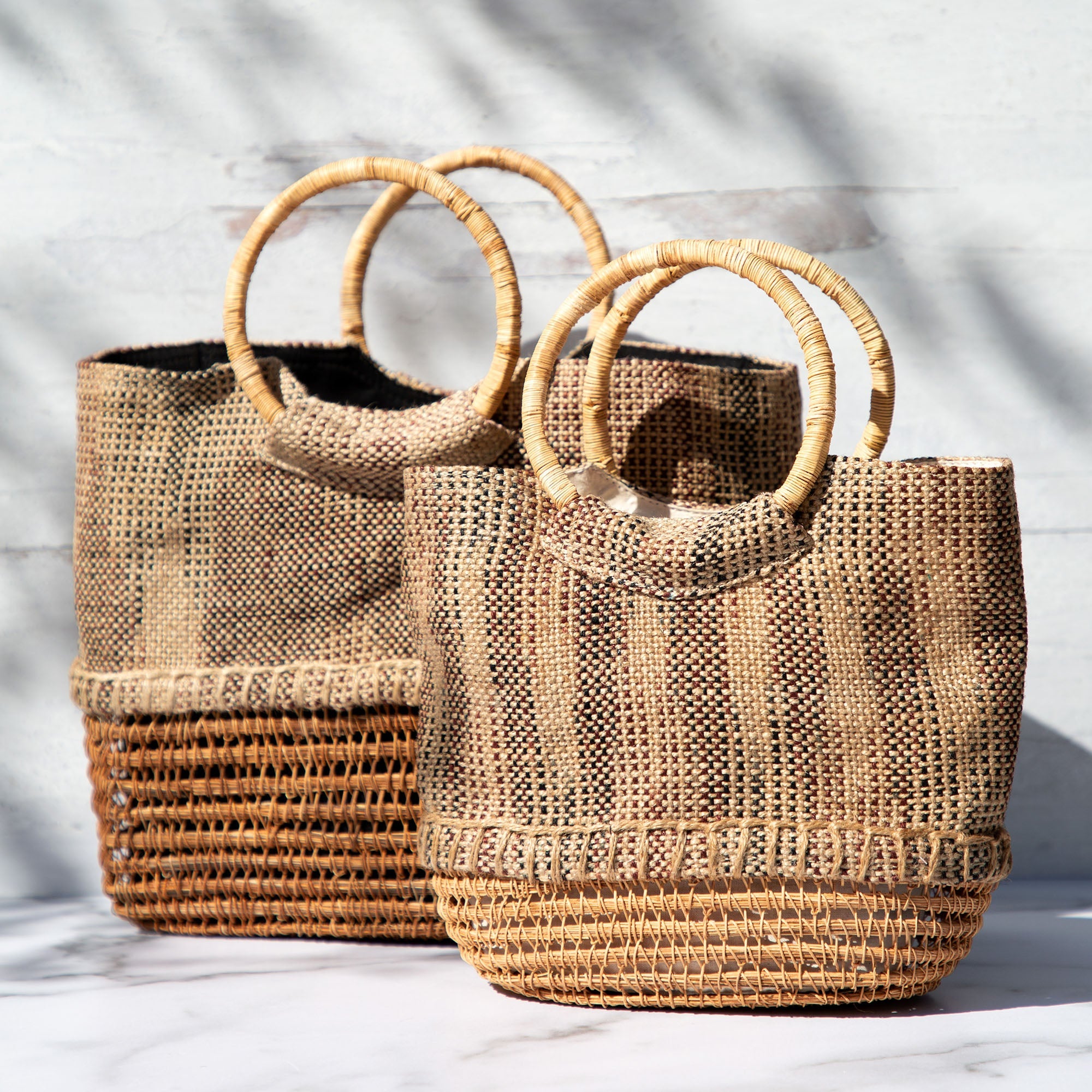 Two palm fiber tote handbags displayed side by side to highlight size options. Handwoven from palm fiber with structured silhouettes and cotton-lined interiors.