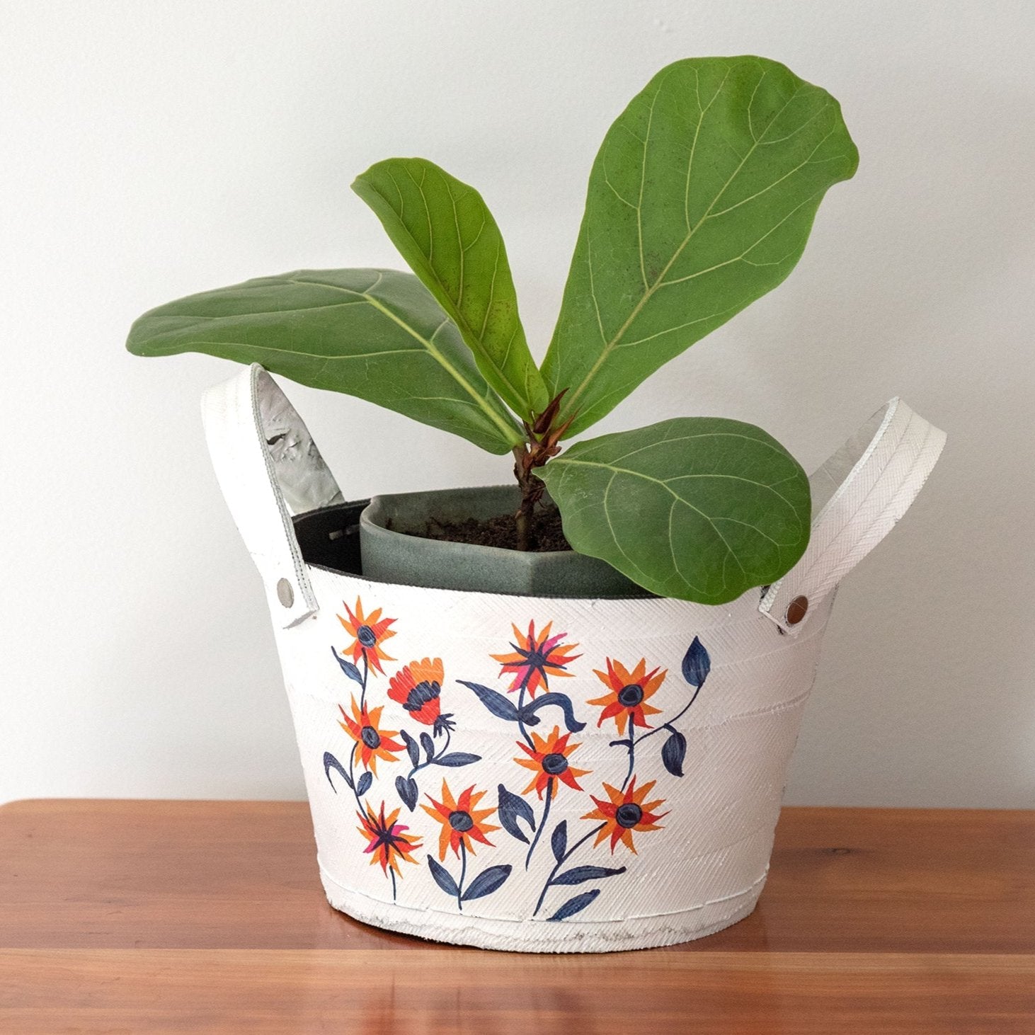 White folklore planter holding a leafy green plant, resting on a light wood surface with a neutral background.