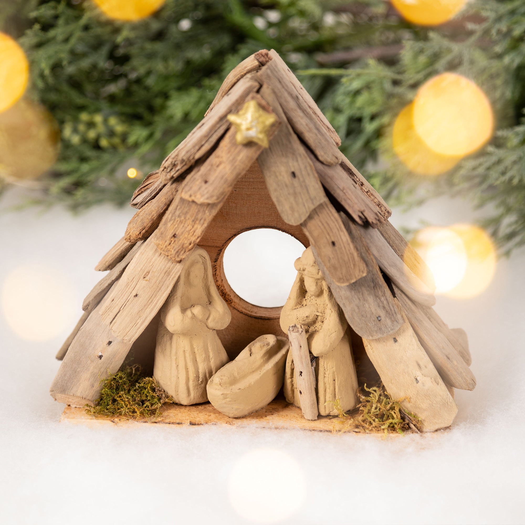 Recycled driftwood nativity ornament with handmade clay figures, featuring Joseph, Mary, and baby Jesus in a rustic stable in front of a Christmas tree.