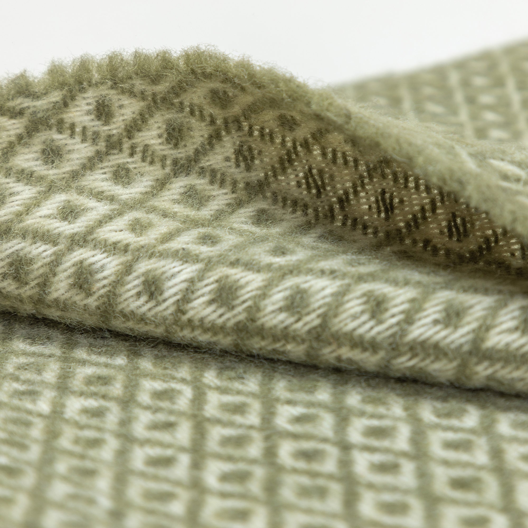 Close-up photo focusing on the fabric texture and pattern of a green blanket with cream fringe, highlighting the woven diamond design.