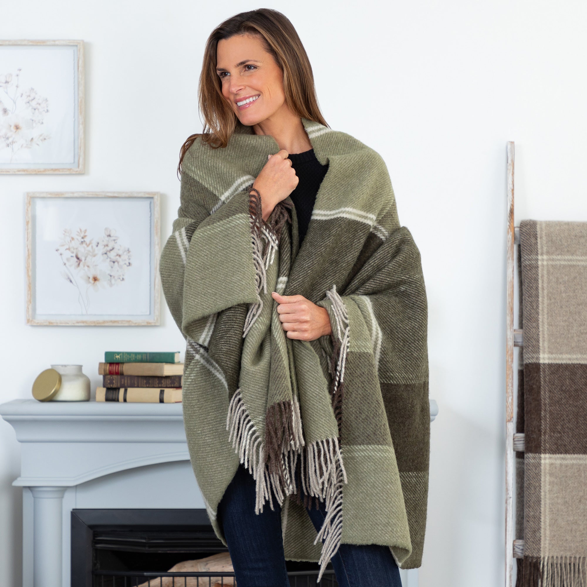 A smiling woman with light brown hair wearing a black sweater, wrapped warmly in a green plaid wool blanket with fringed edges, standing in a cozy room with a gray fireplace, books, and decorative pillows.