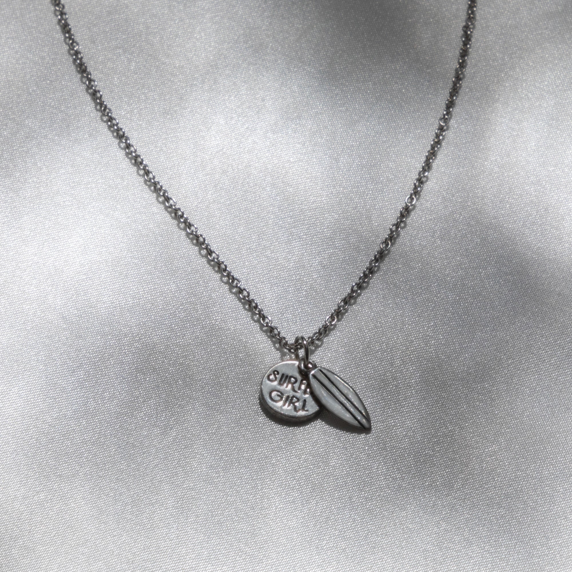 A silver necklace with two small pendants—one circular pendant engraved with "SURF GIRL" and one shaped like a surfboard—resting on a light, shimmery background, celebrating ocean life while supporting plastic cleanup efforts.