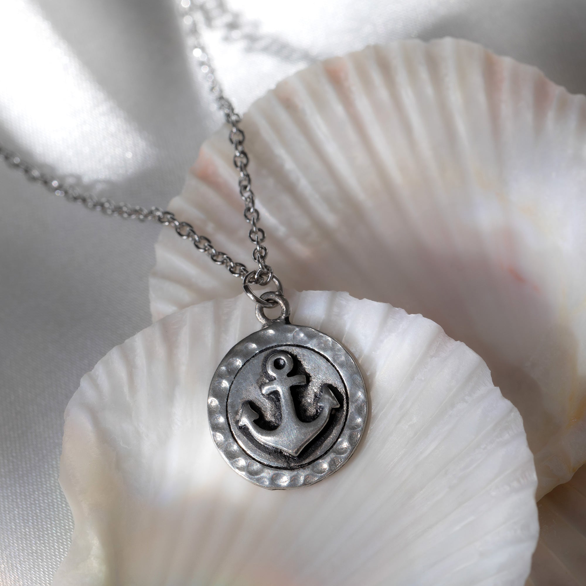 A pewter anchor pendant necklace displayed on white seashells with a soft, light background, symbolizing the ocean’s strength and crafted to support sustainable fashion.