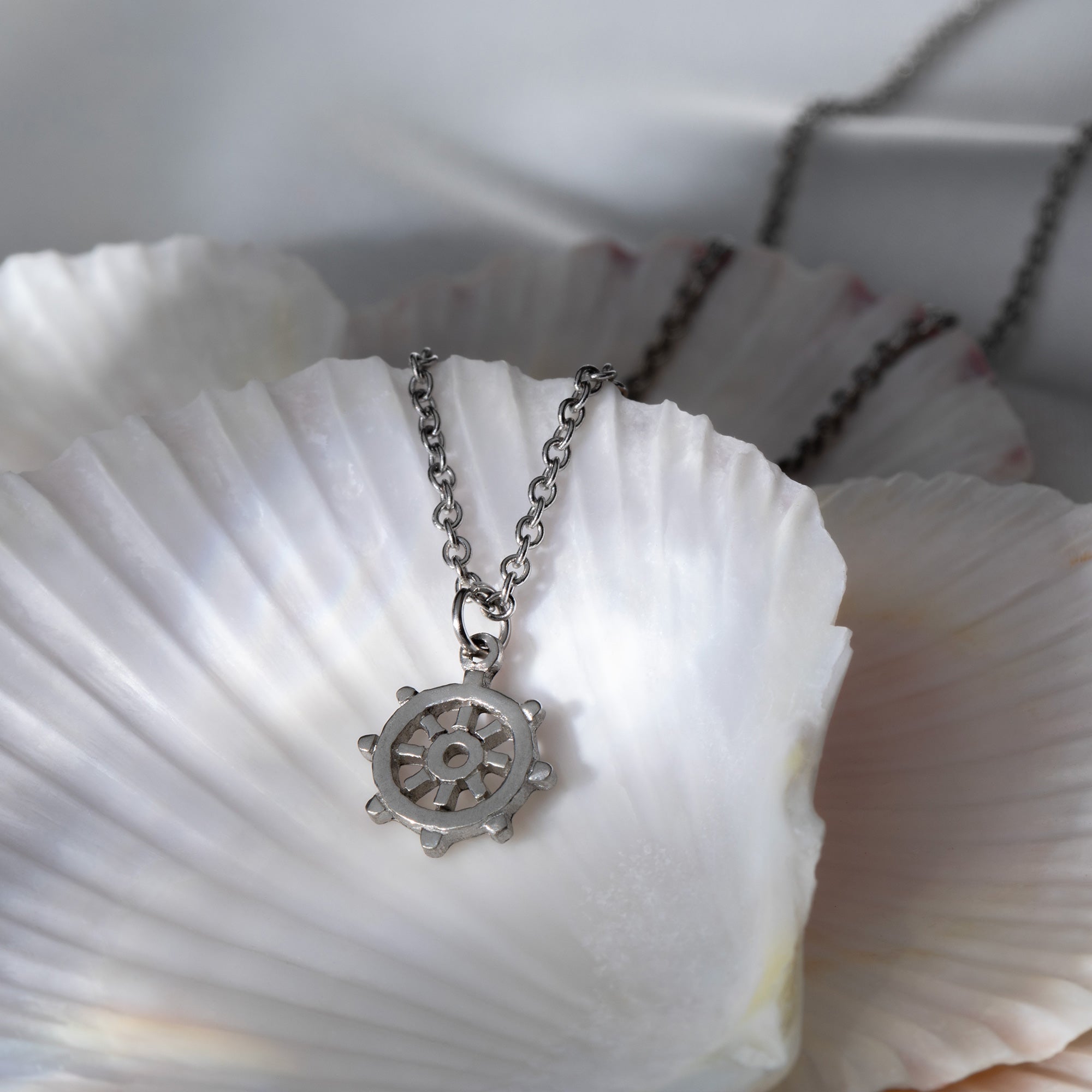 A pewter rudder-shaped pendant necklace nestled inside a seashell, showcasing its intricate design inspired by ocean navigation and the commitment to protecting our seas.