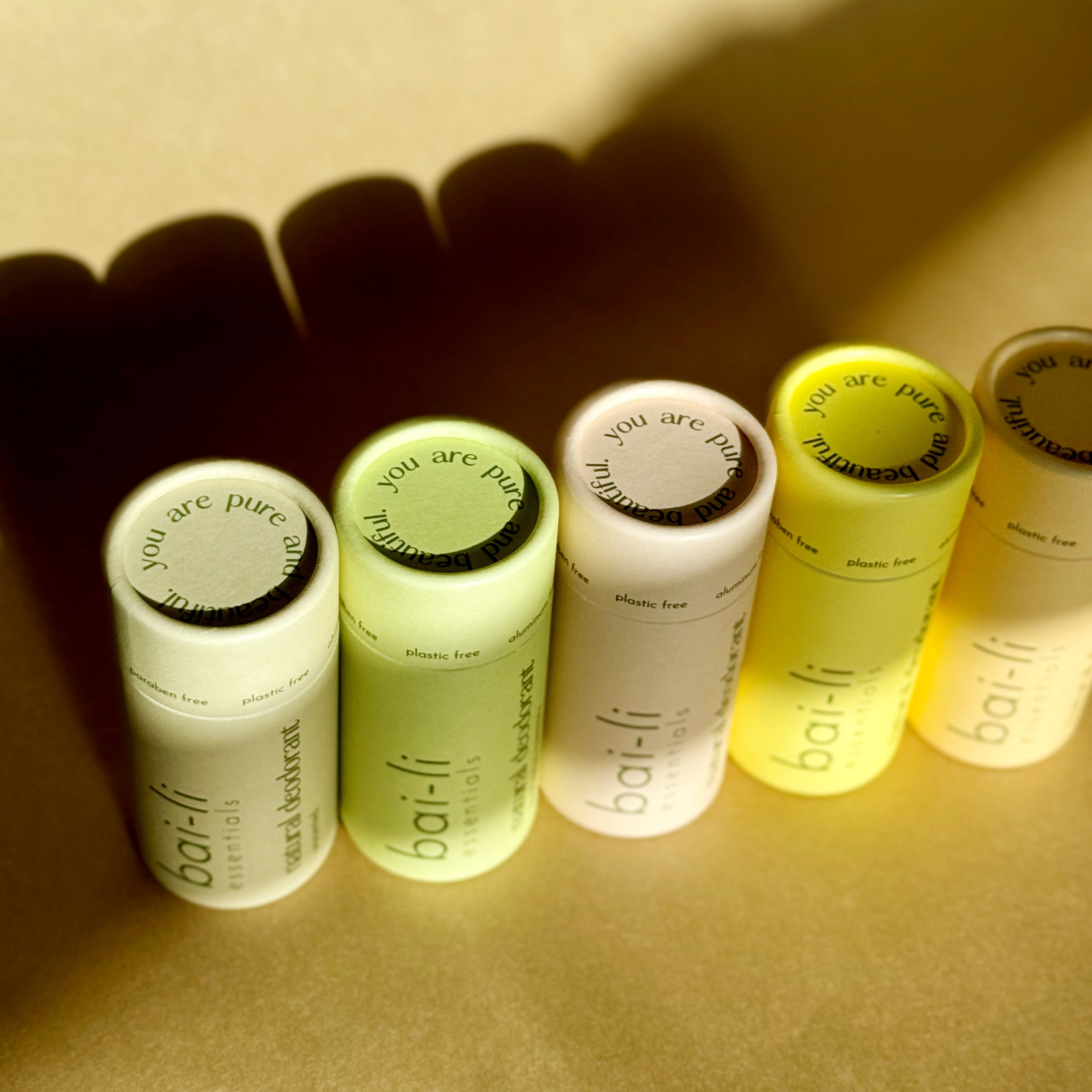 Top view of four plastic-free deodorant tubes in Lavender + Peppermint, Cedarwood + Sweet Orange, Lemon + Bergamot, and Unscented variants placed diagonally on a brown surface.