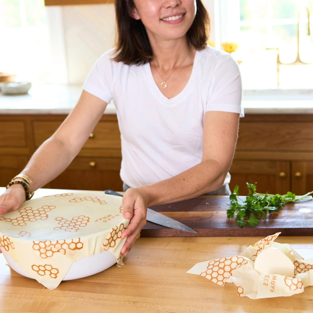 Woman in a white shirt wrapping a bowl with orange honeycomb-patterned reusable beeswax food wrap on a kitchen counter, with a knife and chopped herbs nearby.