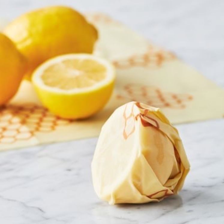 A close-up of a fresh lemon wrapped tightly in beeswax food wrap, with whole and halved lemons blurred in the background on a marble countertop.