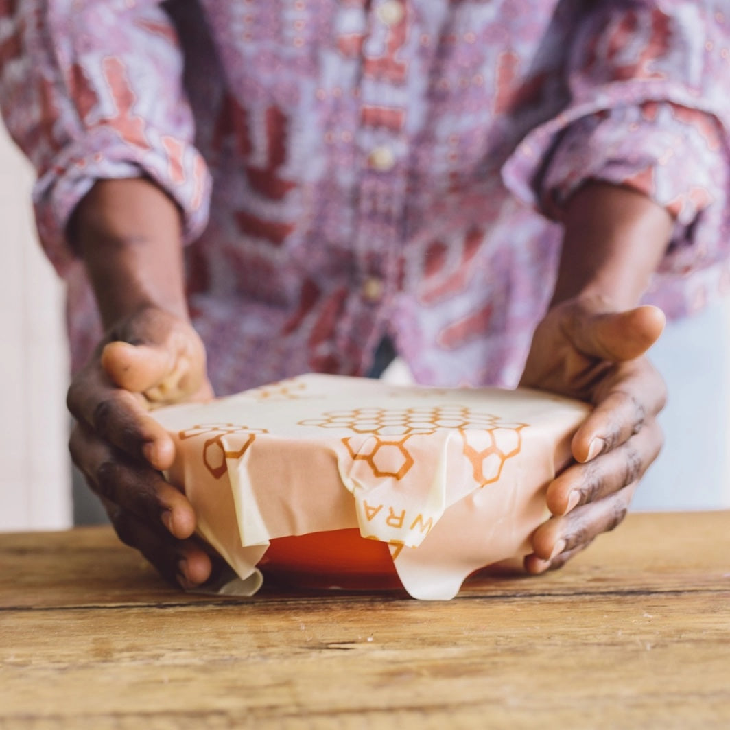 A person carefully covering a bowl with orange-colored beeswax food wrap on a wooden table, with some fresh herbs and a knife nearby.
