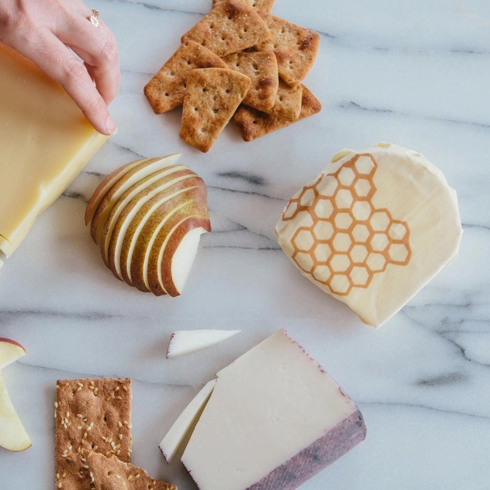 Bees Wrap reusable food wrap with honeycomb design, cheese, crackers, and sliced pear on marble countertop, eco-friendly alternative to plastic.