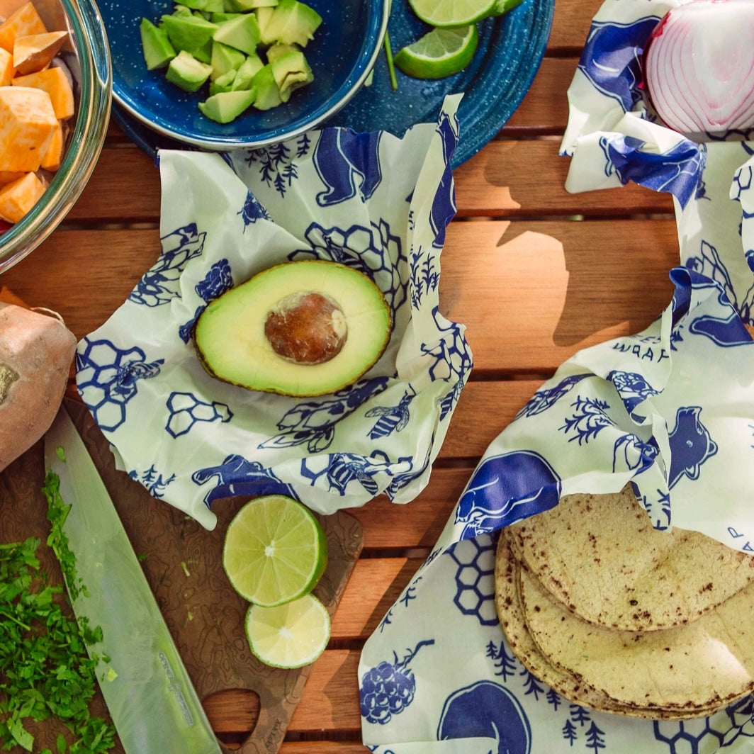 Blue patterned Bees Wrap holding avocado half and tortillas on wooden table with lime slices.