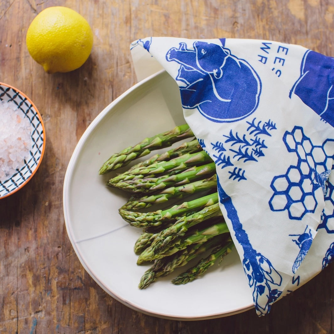 Lemon beside a plate of fresh asparagus partially covered with blue and white patterned beeswax wrap on a wooden table.