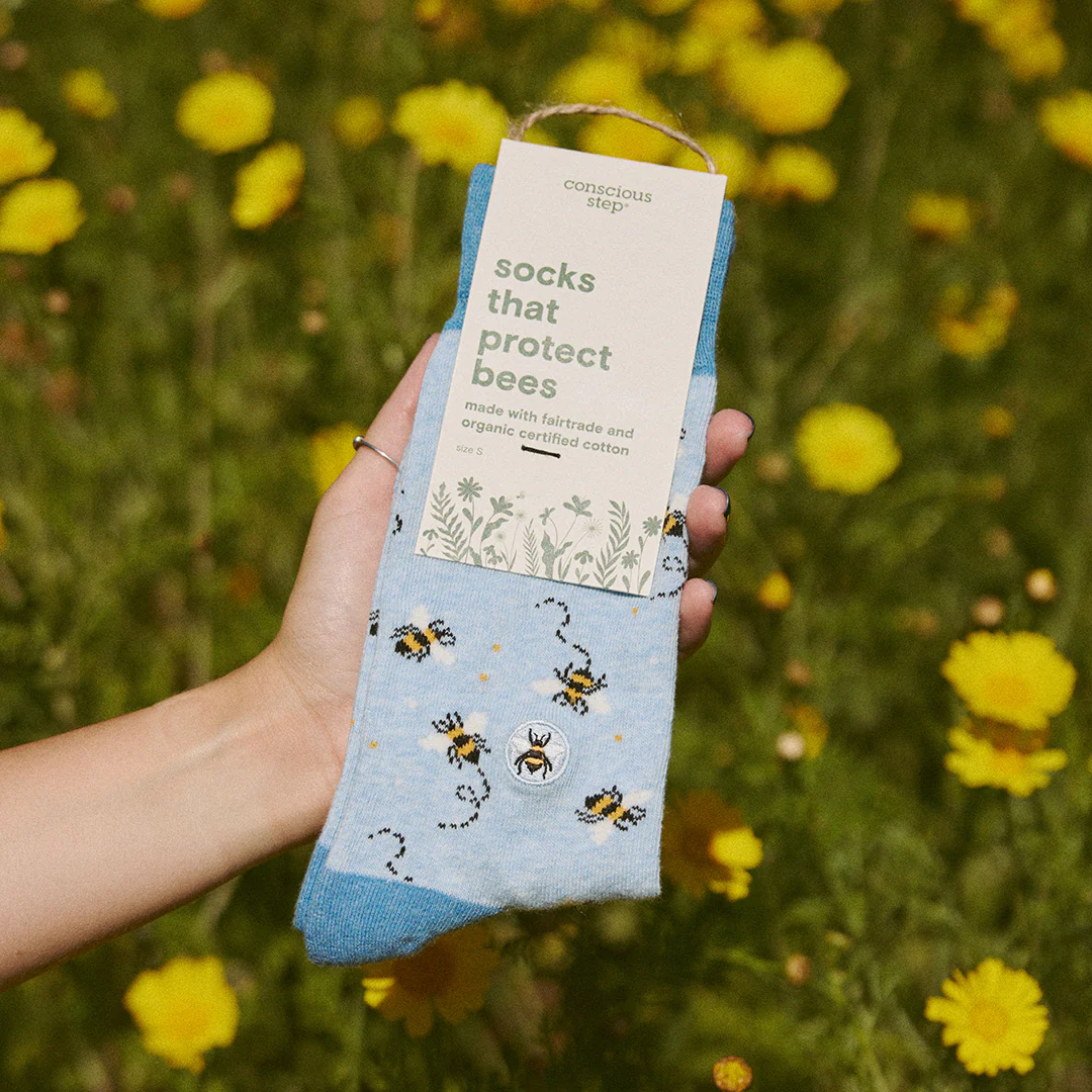 A hand holds a pair of light blue Conscious Step socks with a bee and dotted line pattern against a background of blurred yellow flowers. The tag identifies them as "socks that protect bees" and states they are made with fairtrade and organic certified cotton.