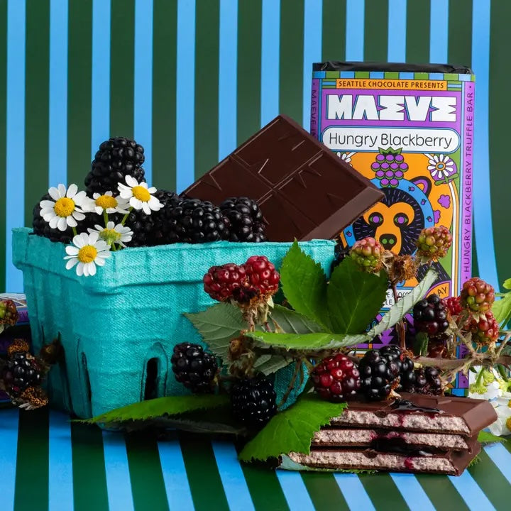 A vibrant turquoise basket filled with fresh blackberries and white daisies, accompanied by a bar of Maeve fair trade chocolate labeled "Hungry Blackberry."