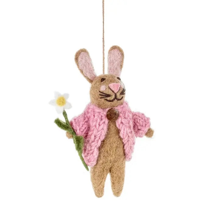 A handmade felt rabbit is shown, wearing a pink knit cardigan with a brown button. The rabbit is holding a white flower with a yellow center and green stem.