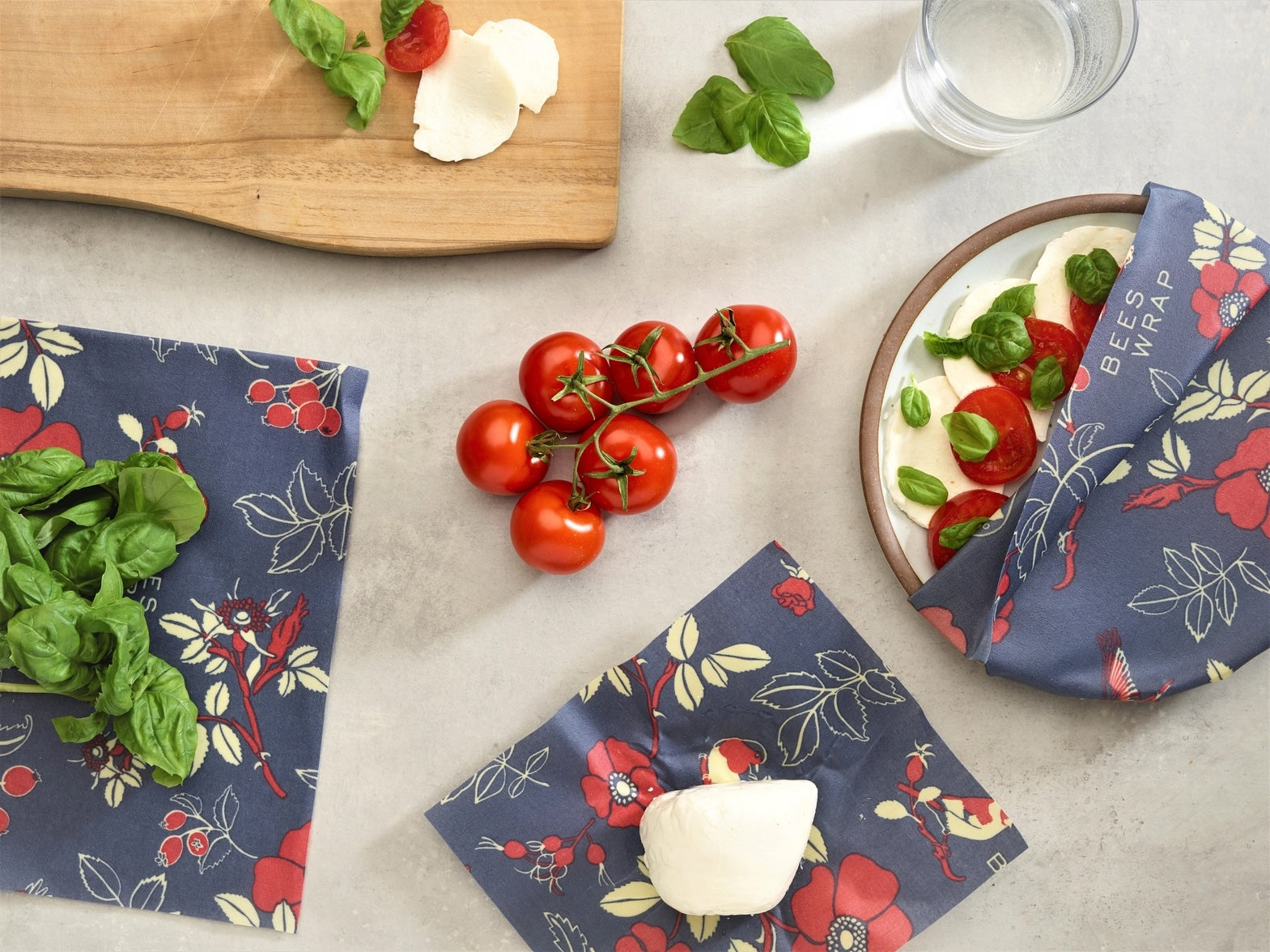 Three Bees Wraps with blue background and red floral and berry patterns, one wrap partially covering a plate of tomato, basil, and mozzarella, with a glass of water nearby.