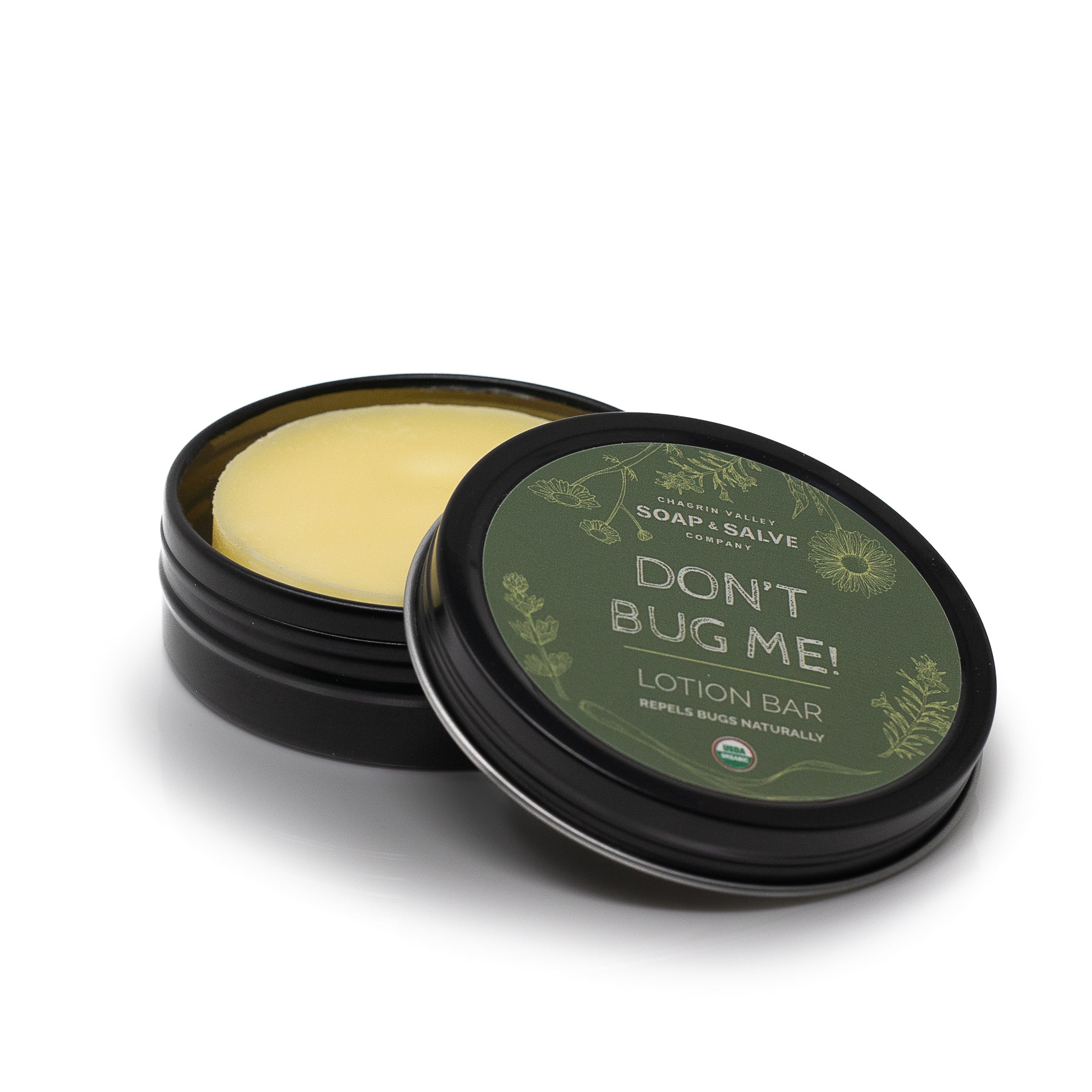 A 'Don't Bug Me!' natural repellent lotion bar in a small, round black tin. The lid is open, showing a yellow lotion bar inside and the green label on the lid with decorative drawings of leaves.