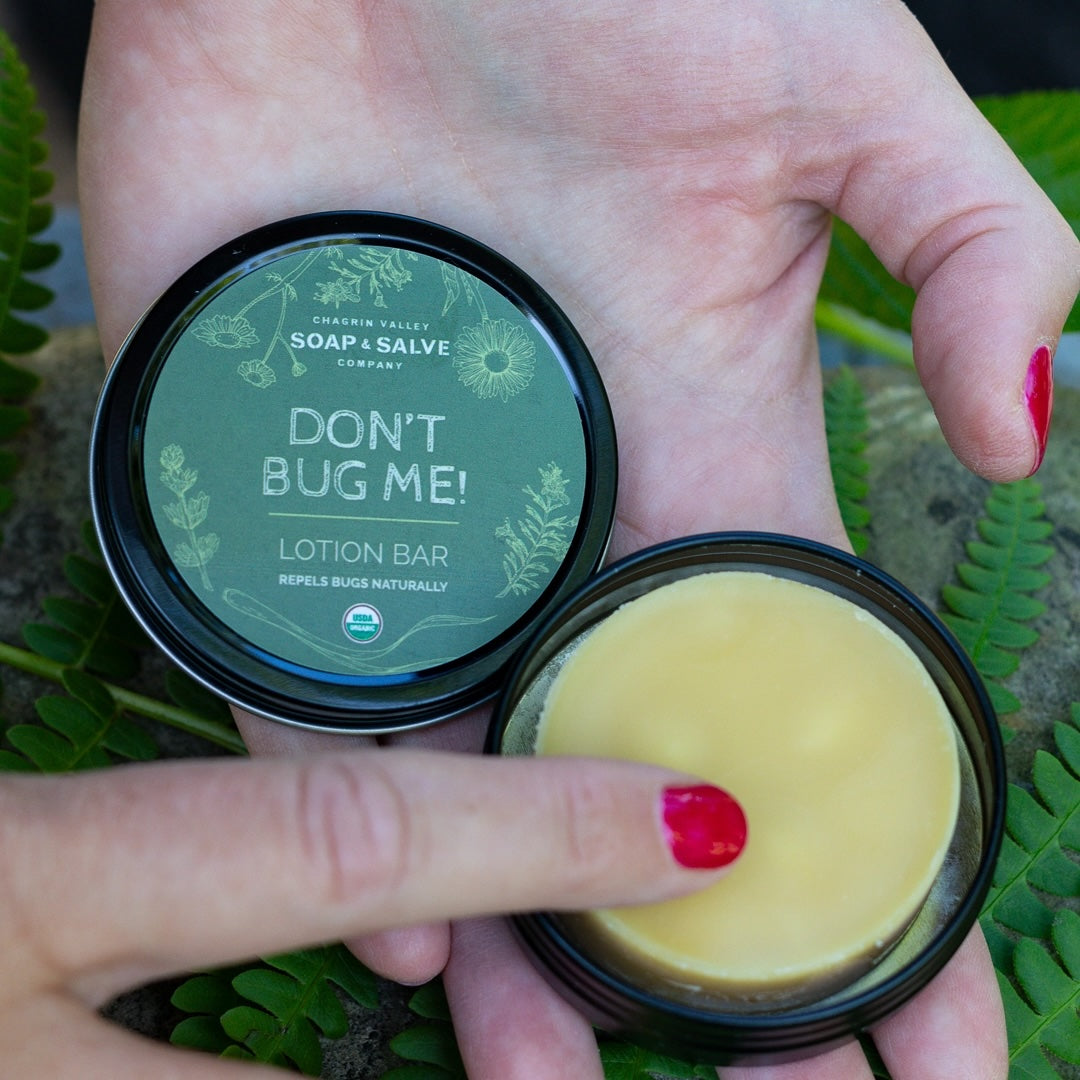 A pair of hands holds an open tin of 'Don't Bug Me!' natural bug repellent lotion bar. A finger touches the yellow lotion bar displaying the ergonomic proportions and use of the product.