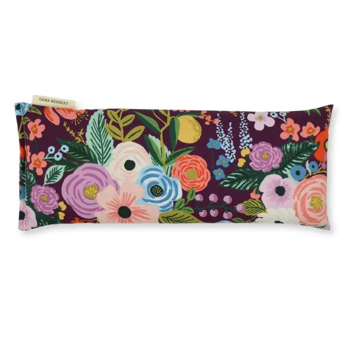 A rectangular eye pillow by Dana Herbert has a vibrant floral pattern with purple pink and blue flowers on a dark purple background.